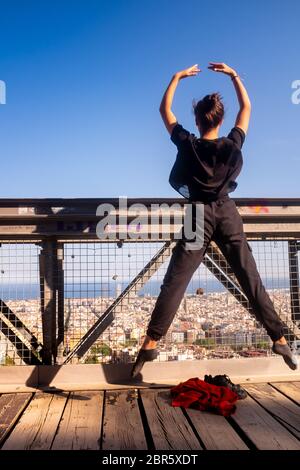 Elegant ballet performance by a young female dancer wearing a black dress, she is jumping in mid-air in front of a panoramic view of Barcelona, Spain.