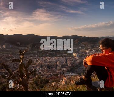 Sad girl sitting on the hill embracing her knees and looking away at sunset, sadly depressed teenager spending time alone, thoughtful young girl feeli Stock Photo