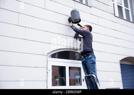 Young Serviceman Standing On Ladder Checking Outdoor Electric Lamp On Wall With Torch Light Stock Photo