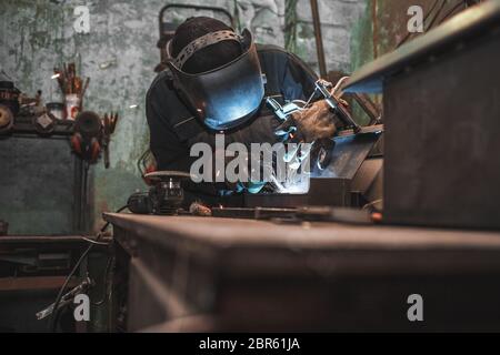 Worker in a welder mask works in a workshop for welding iron. Man makes iron products. Guy works with a welding machine using a mask to protect his ey Stock Photo