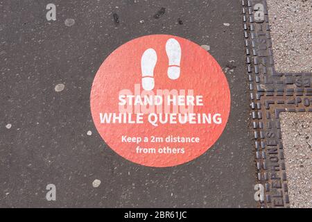 Social distancing queue sign - stand here while queueing' sign on pavement during coronavirus pandemic - UK Stock Photo