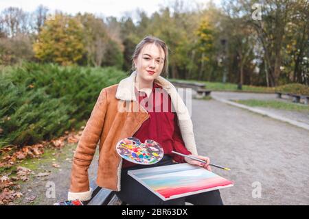 Portrait of a street artist who sits in a park on a bench and holds a painting, palette and brush in her hands Stock Photo