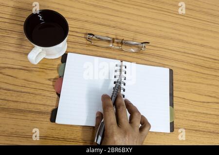 Businessman writing in his diary. Open notebook with blank pages next to cup of coffee and eyeglasses on wooden table. Business still life concept wit Stock Photo