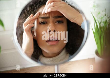 Depressed african teen girl squish pimple looking in mirror at table. Stock Photo