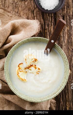 Fresh homemade cream of cauliflower soup garnished with roasted cauliflower floret slices, served in bowl with spoon, photographed overhead on rustic Stock Photo