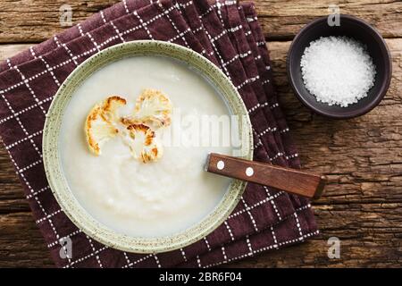 Fresh homemade cream of cauliflower soup garnished with roasted cauliflower floret slices, served in bowl with spoon, salt on the side, photographed o Stock Photo