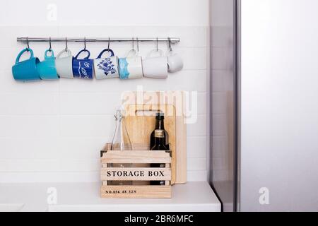 Multi-colored ceramic cups in blue tones hang on hooks above the kitchen table. Bottles with olive oil stand in a wooden box. Stock Photo