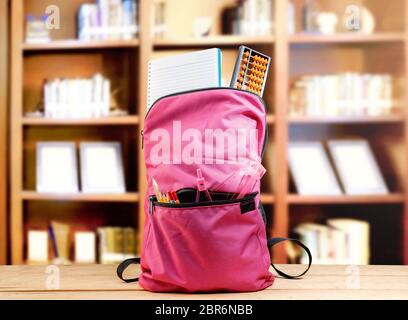 Pink backpack with book and different stationery on the wooden table in library. Back to School concept Stock Photo