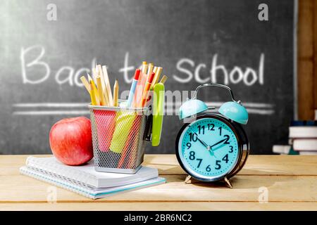 Pile of notebooks with apple and pencils in basket container with green stapler and alarm clock on the wooden table with blackboard background. Back t Stock Photo