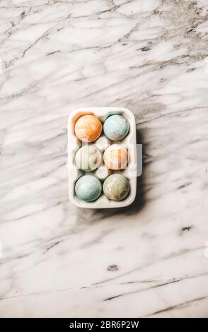 Flat-lay of colorful pastel dyed Easter eggs in white ceramic holder over white marble background, top view Stock Photo