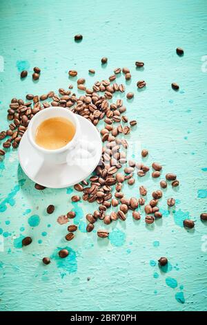 Freshly brewed small white generic cup of espresso coffee with roasted beans scattered over a painted texture blue background with copy space Stock Photo