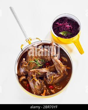 Leg portions of wild rabbit venison in a savory sauce seasoned with orange zest and herbs viewed top down on white with a pot of red cabbage as sides Stock Photo