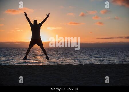A rear-view shot of an unrecognizable man doing jumping jacks on the beach, he is looking out towards the sea and the sunset on a beautiful evening in Stock Photo