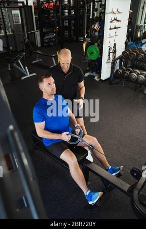 A  high-angle view of a mid-adult caucasian man training in the gym, he is using a rowing machine. A fitness instructor can be seen standing by the ma Stock Photo