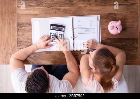 An Overhead View Of Couple Calculating Tax Invoice Using Calculator On Wooden Desk At Home Stock Photo
