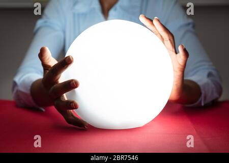 Close-up Of Fortuneteller's Hand Covering The Glowing Crystal Ball On Red Desk Stock Photo