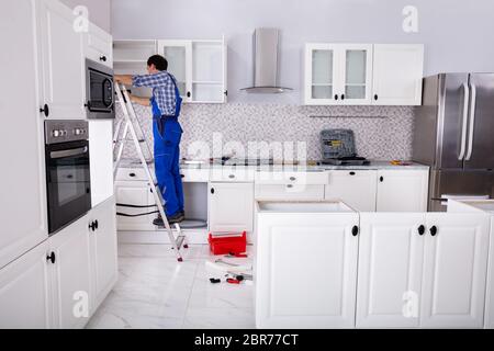 Rear View Of A Man Standing On Ladder Adjusting Cabinet Door Hinge On Kitchen Cabinet Stock Photo