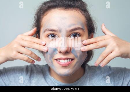 Cute and happy girl smears face with cosmetic clay or mud and smiles. Cosmetic mask, face scrub. The concept of health and beauty. Stock Photo