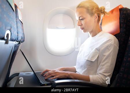 Attractive caucasian female passenger working at modern laptop computer using wireless connection on board of commercial airplane flight. Stock Photo
