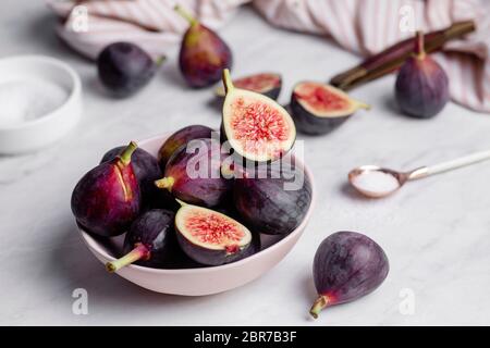 Close up of a bowl of ripe, purple figs on a white marble countertop surrounded by a striped napkin, and a rose gold spoonful of sugar Stock Photo