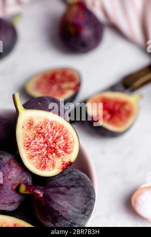 Close up of a halved fig, sitting on top of other purple figs in a bowl with a knife, striped napkin, and white marble countertop in the blurred backg Stock Photo