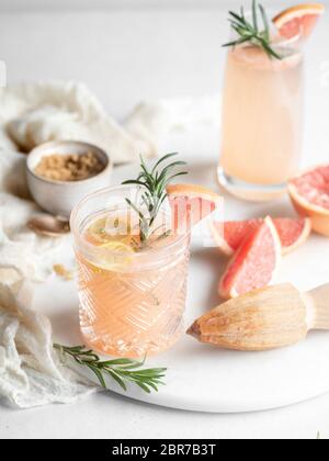 Old fashioned drinking glass filled with a grapefruit juice cocktail and garnished with lemon and rosemary on a white tray with a wooden hand juicer Stock Photo