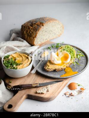 A side shot of a slice of bread topped with hummus, herbs, and a soft boiled egg, on a silver tray and wood cutting board on a white marble countertop Stock Photo