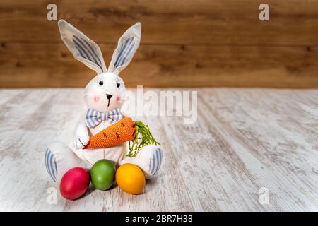 Close-up of three colorful Easter eggs in front of Easter bunny holding carrot on white painted and vintage oak background, front view Stock Photo