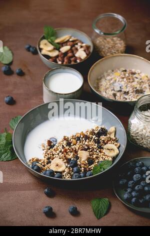 Homemade crunchy puffed millet grain granola with dried fruits and nuts in ceramic bowl, with yogurt, mint and ingredients above. Brown texture backgr Stock Photo