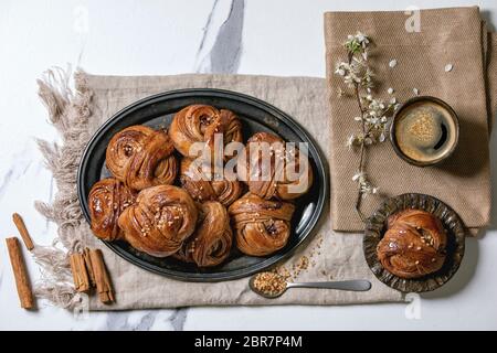 Traditional Swedish cinnamon sweet buns Kanelbulle on vintage tray, cup of coffee, cinnamon sticks, blossom branches on linen cloth over white marble Stock Photo