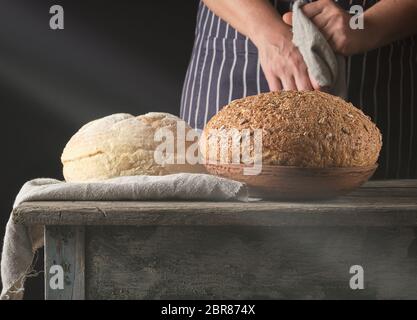 ray of light falls on baked round bread on a table and a woman in a striped apron, black background, concept of cooking Stock Photo