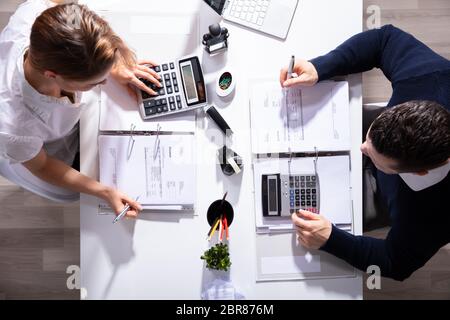 High Angle View Of Male And Female Businesspeople Calculating Invoice With Calculator In Office Stock Photo