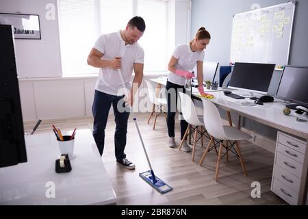 Two Smiling Young Janitor Cleaning The Desk And Mopping Floor In The Office Stock Photo