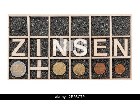 Plywood letters arranged in a collecting box with black decorative sand depicting the word ZINSEN and some Euro coins a nearby subjects in top view is Stock Photo