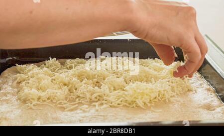 Pizza dough or bread dough and grated Mozzarella cheese close up on baking sheet. Woman putting cheese on dough Stock Photo