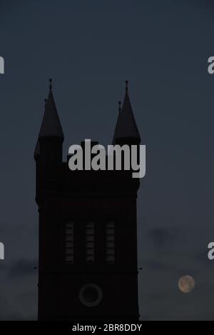 A setting full moon sits in the early morning dark blue sky next to the silhouetted church tower of St. Andrew's Presbyterian Church in Victoria, Brit Stock Photo