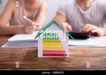 Close-up Of A House Showing Energy Efficiency Rate In Front Of Couple Using Calculator Stock Photo