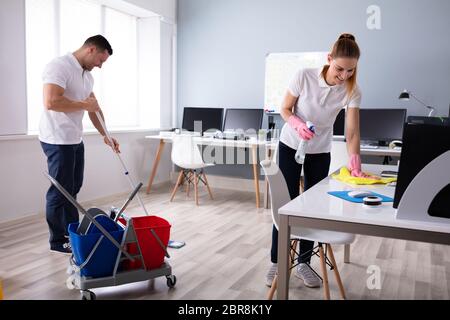 Two Smiling Young Janitor Cleaning The Desk And Mopping Floor In The Office Stock Photo