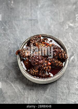 Delicious jam with baby pine cones in small bowl. Traditional Siberian dessert - young pine cones jam on gray textured background. Top view or flat la Stock Photo