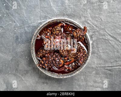 Delicious jam with baby pine cones in small bowl. Traditional Siberian dessert - young pine cones jam on gray textured background. Top view or flat la Stock Photo