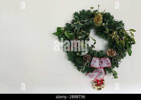 Home made Christmas wreath with holly, mistletoe, pine cones, ivy, tinsel, ribbon, artficial conifer twigs and bells against a light background with c Stock Photo