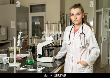 Young, Caucasian white, female laboratory technician in white robe with stethoscope standing next to compound microscope, laboratory, equipment and gl Stock Photo