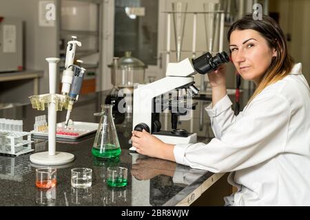 Middle age, Caucasian white, female laboratory technician in white robe with stethoscope sitting next to compound microscope, laboratory, equipment an Stock Photo