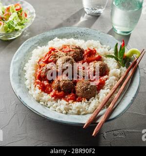 Overhead view on bowl of tasty asian rice with large saucy meatballs and chopsticks on rim of bowl next to water glass Stock Photo