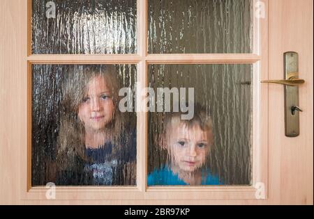 Portrait of a cute little Caucasian boy and girl hiding behind a door with glass windows while playing hide and seek Stock Photo