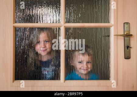 Portrait of a cute little Caucasian boy and girl hiding behind a door with glass windows while playing hide and seek Stock Photo