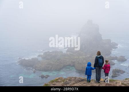 Mother with her children standing on the top of a cliff looking at the rock in the sea at the Lands End - the most westerly point of England, Penwith Stock Photo