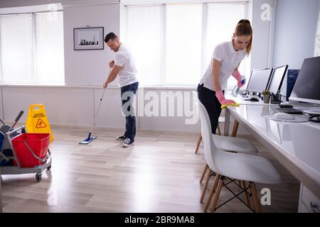 Two Smiling Young Janitor Cleaning The Desk And Mopping Floor In The Office Stock Photo