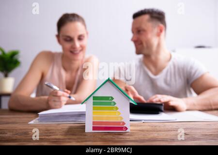 House Showing Energy Efficiency Rate In Front Of Happy Couple Using Calculator Over Wooden Desk Stock Photo