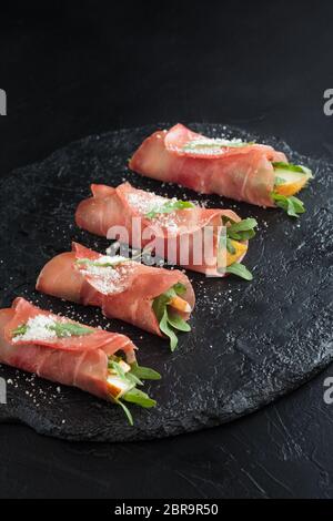 Prosciutto rolls stuffed with arugula and pears on black slate. Stock Photo